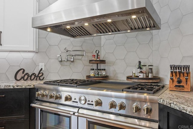 kitchen featuring range with two ovens, light stone countertops, dark cabinetry, decorative backsplash, and wall chimney exhaust hood