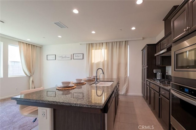 kitchen with stainless steel appliances, a kitchen island with sink, a sink, dark brown cabinets, and under cabinet range hood