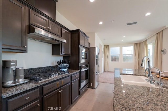 kitchen featuring dark brown cabinetry, dark stone countertops, stainless steel appliances, under cabinet range hood, and a sink