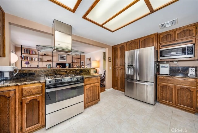 kitchen with island range hood, visible vents, appliances with stainless steel finishes, dark stone counters, and brown cabinetry