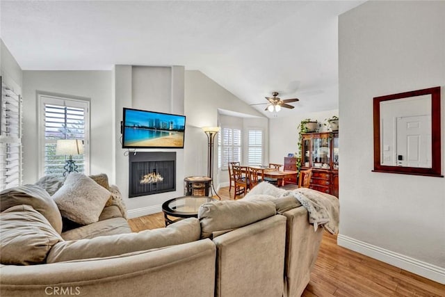 living room featuring vaulted ceiling, light wood-style flooring, baseboards, and a warm lit fireplace