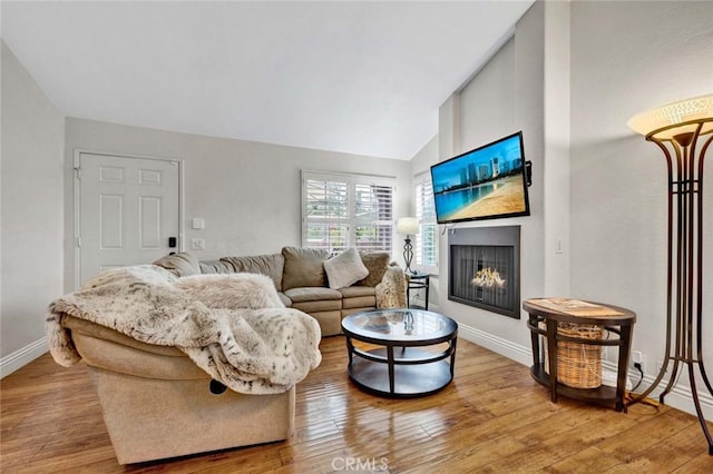 living area featuring a warm lit fireplace, light wood-type flooring, baseboards, and vaulted ceiling