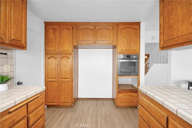 kitchen featuring tasteful backsplash, tile countertops, stainless steel oven, light wood-style flooring, and brown cabinetry