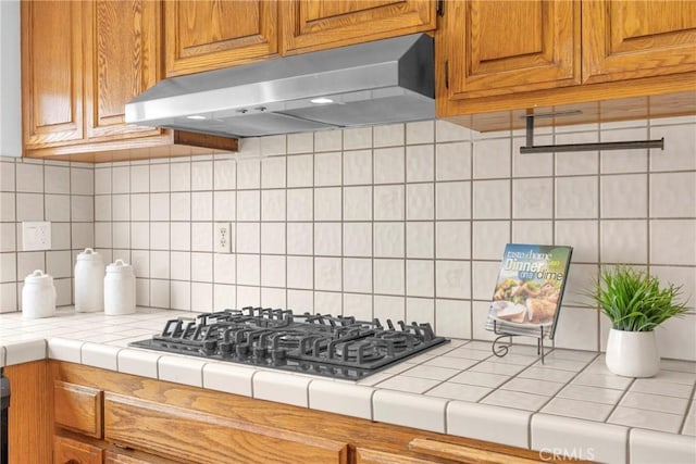 kitchen with black gas stovetop, under cabinet range hood, tile countertops, brown cabinetry, and decorative backsplash