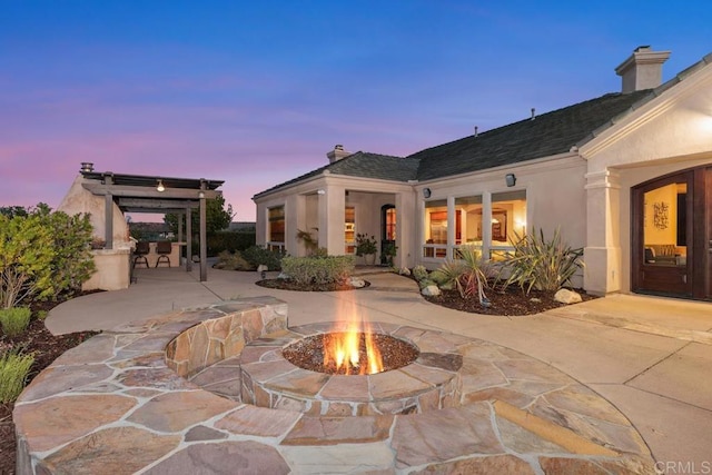 back of property at dusk featuring a chimney, a patio, a fire pit, and stucco siding