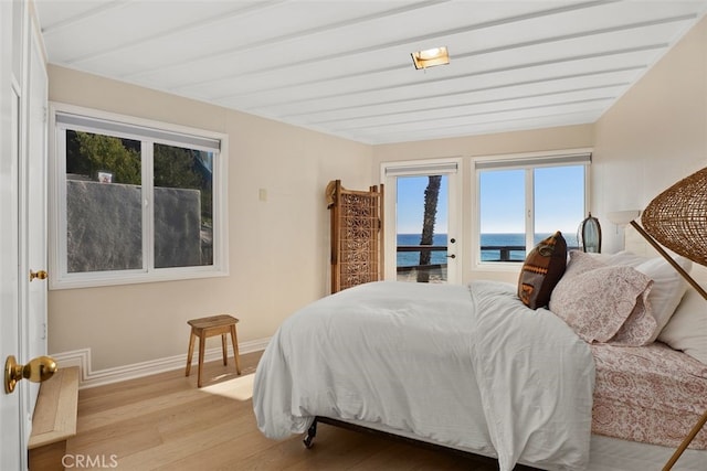 bedroom featuring baseboards, a water view, access to exterior, light wood-style floors, and beam ceiling