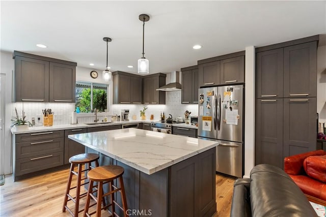 kitchen featuring light wood-style flooring, stainless steel appliances, a sink, a kitchen breakfast bar, and wall chimney exhaust hood