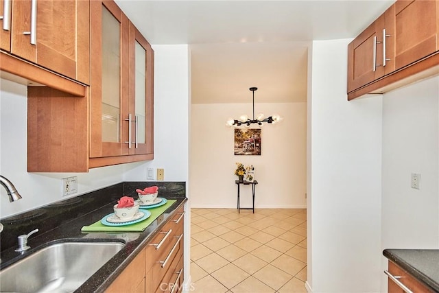 kitchen with light tile patterned floors, a sink, brown cabinetry, dark stone countertops, and glass insert cabinets