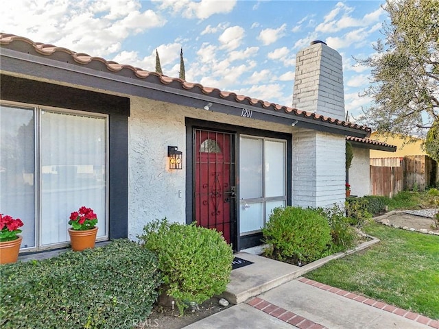 property entrance featuring a tiled roof, a chimney, fence, and stucco siding