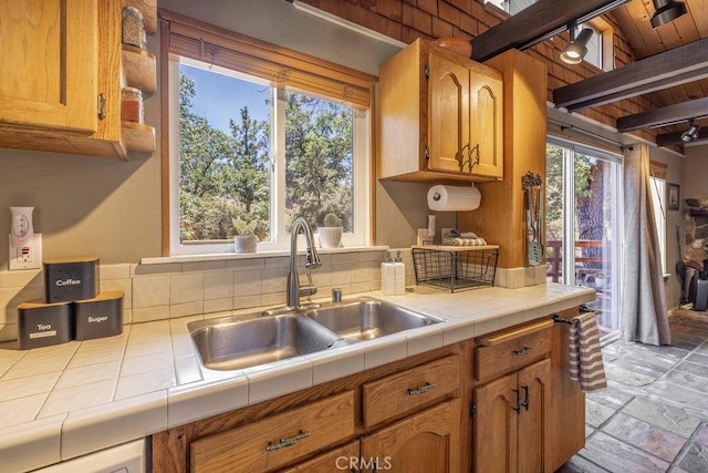 kitchen with beam ceiling, brown cabinets, stone tile floors, tile counters, and a sink