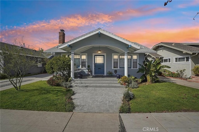 bungalow-style house featuring driveway, covered porch, a chimney, and a lawn