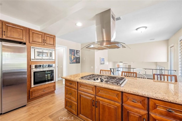 kitchen with light wood finished floors, island range hood, light stone counters, brown cabinets, and stainless steel appliances