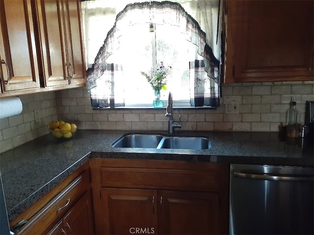 kitchen with dark countertops, backsplash, stainless steel dishwasher, brown cabinetry, and a sink