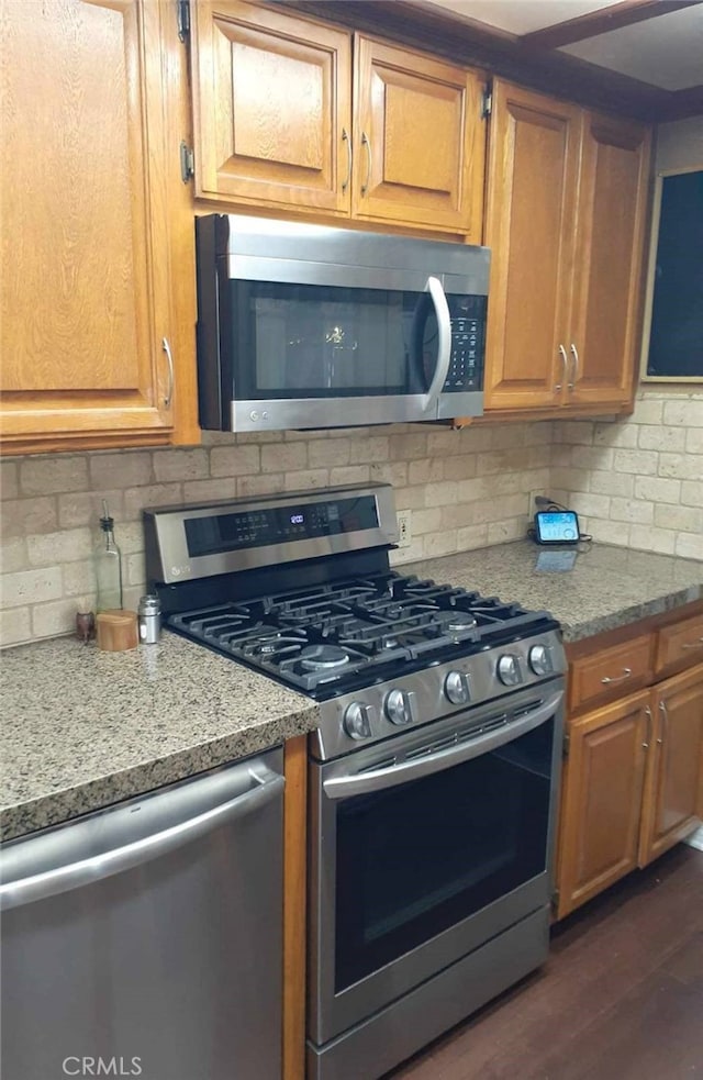 kitchen with light stone countertops, appliances with stainless steel finishes, brown cabinets, dark wood-style floors, and tasteful backsplash