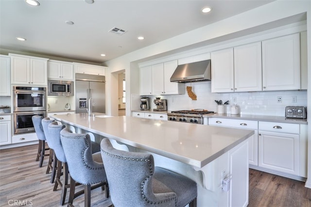 kitchen featuring wall chimney exhaust hood, a center island with sink, built in appliances, visible vents, and wood finished floors