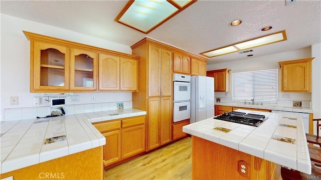 kitchen with a textured ceiling, light wood-style flooring, white appliances, tile counters, and glass insert cabinets