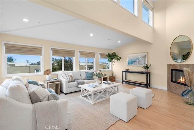 living area with baseboards, a glass covered fireplace, a towering ceiling, light wood-type flooring, and recessed lighting