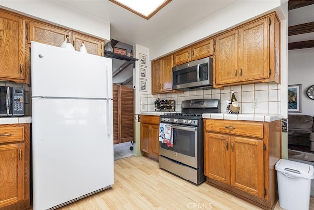 kitchen featuring stainless steel appliances, tile counters, backsplash, brown cabinetry, and light wood-type flooring