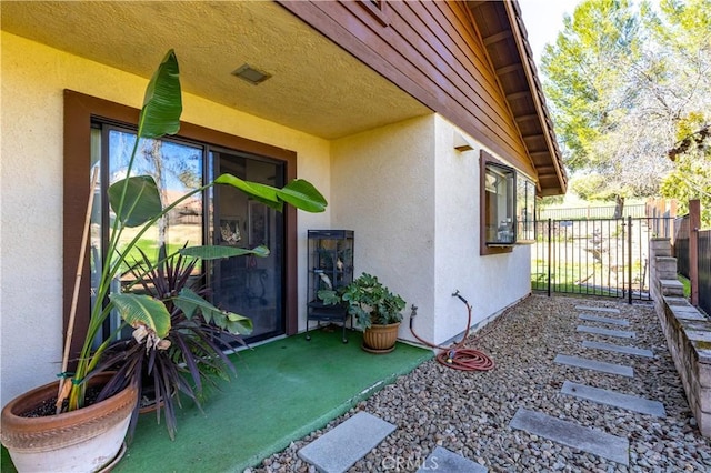 view of exterior entry featuring visible vents, a gate, fence, and stucco siding