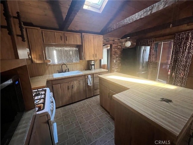 kitchen featuring white gas stove, brown cabinetry, freestanding refrigerator, lofted ceiling with skylight, and a sink