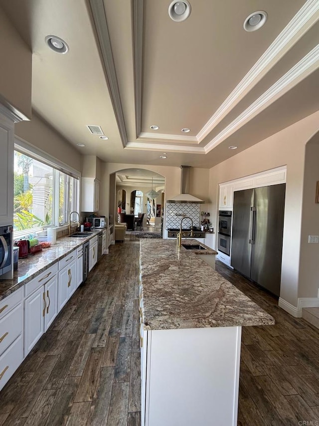 kitchen with arched walkways, a raised ceiling, appliances with stainless steel finishes, white cabinets, and wall chimney exhaust hood