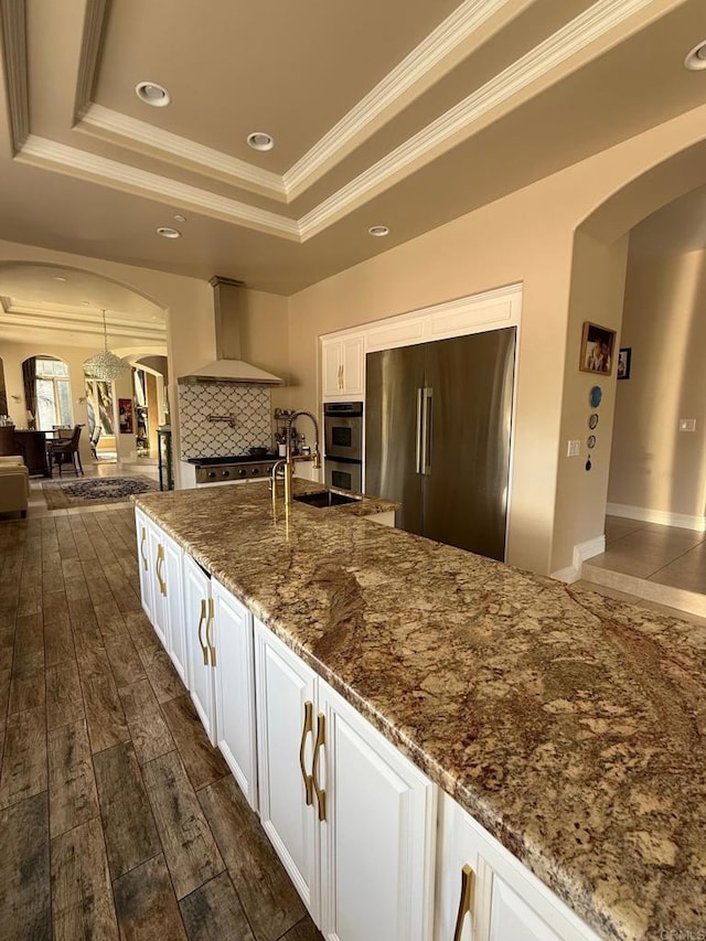 kitchen featuring arched walkways, wall chimney exhaust hood, a tray ceiling, and high end refrigerator