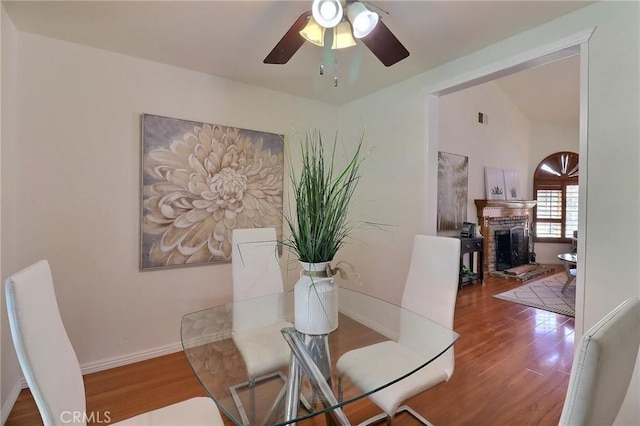 dining space with a fireplace with raised hearth, ceiling fan, baseboards, and wood finished floors