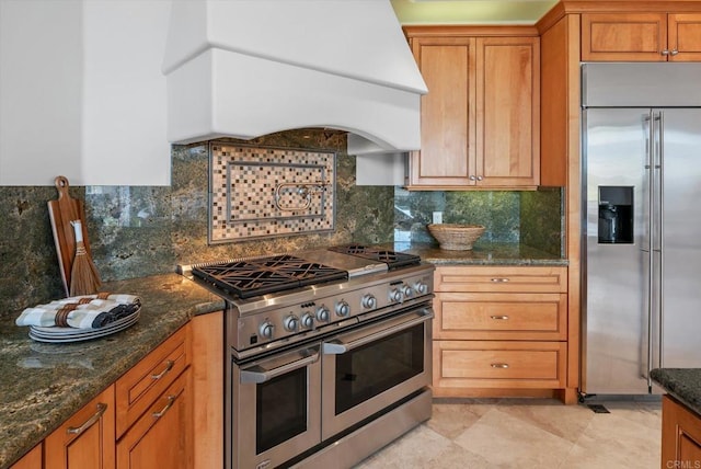 kitchen with premium appliances, brown cabinets, decorative backsplash, dark stone counters, and custom range hood