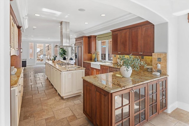 kitchen with stone tile flooring, ornamental molding, glass insert cabinets, island range hood, and baseboards