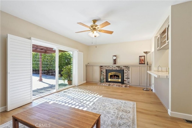 living room featuring baseboards, light wood finished floors, ceiling fan, and a brick fireplace