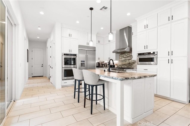 kitchen featuring a kitchen island with sink, stainless steel appliances, a sink, visible vents, and wall chimney range hood