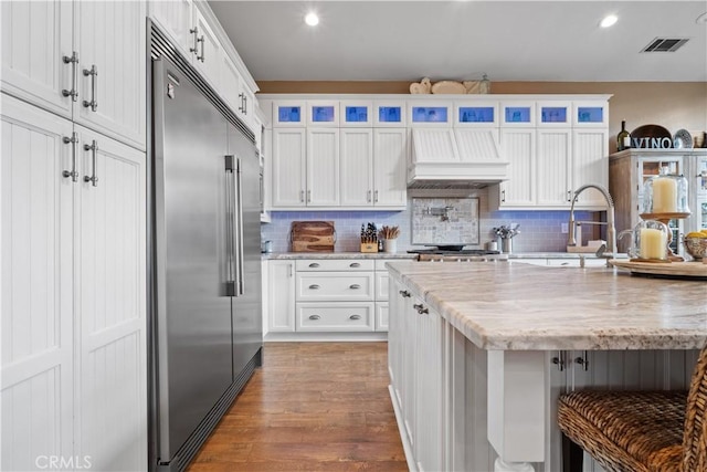 kitchen featuring premium range hood, wood finished floors, visible vents, stainless steel built in fridge, and tasteful backsplash