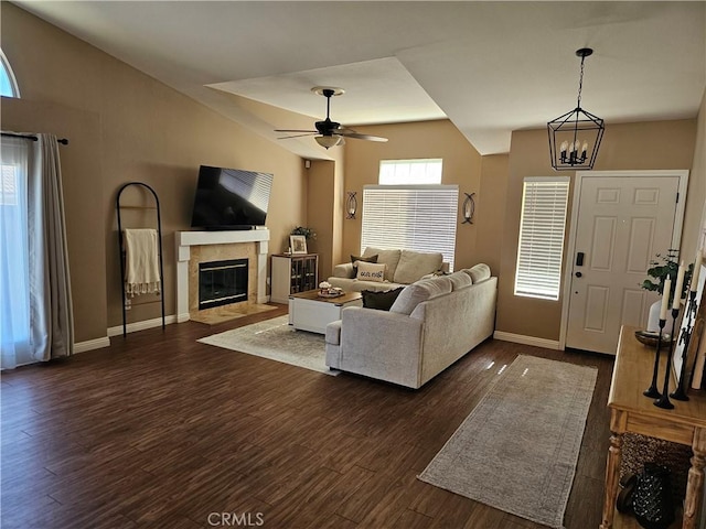 living room with lofted ceiling, ceiling fan with notable chandelier, dark wood-style flooring, a fireplace with flush hearth, and baseboards