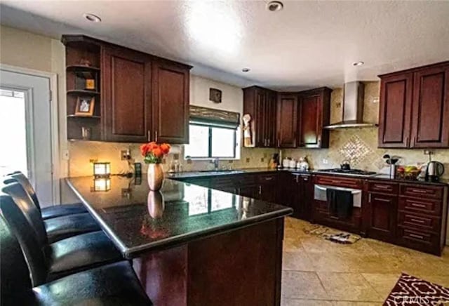 kitchen featuring wall chimney exhaust hood, open shelves, gas cooktop, a peninsula, and a kitchen bar