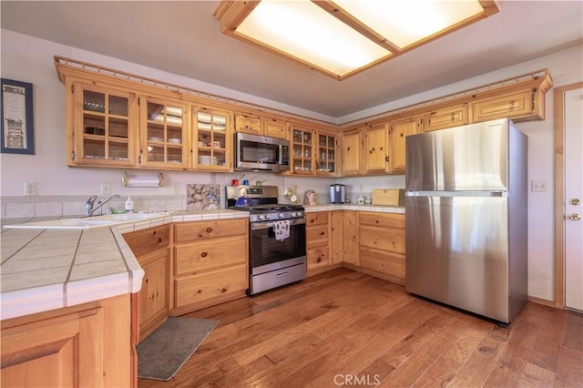 kitchen featuring stainless steel appliances, tile counters, glass insert cabinets, light wood-style floors, and a sink