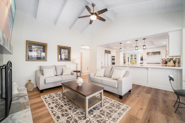 living area featuring a ceiling fan, beam ceiling, a fireplace with raised hearth, arched walkways, and light wood-type flooring