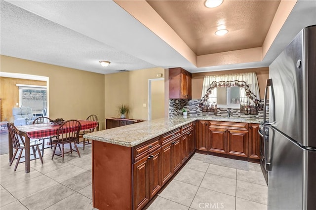 kitchen featuring light tile patterned floors, a peninsula, backsplash, freestanding refrigerator, and light stone countertops