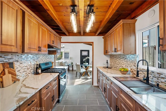 kitchen featuring stainless steel gas range oven, light stone counters, a sink, under cabinet range hood, and beam ceiling