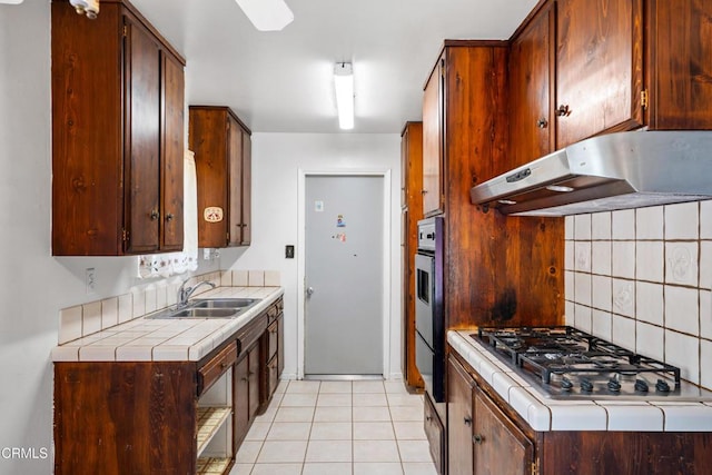 kitchen with under cabinet range hood, stainless steel gas cooktop, a sink, backsplash, and tile counters