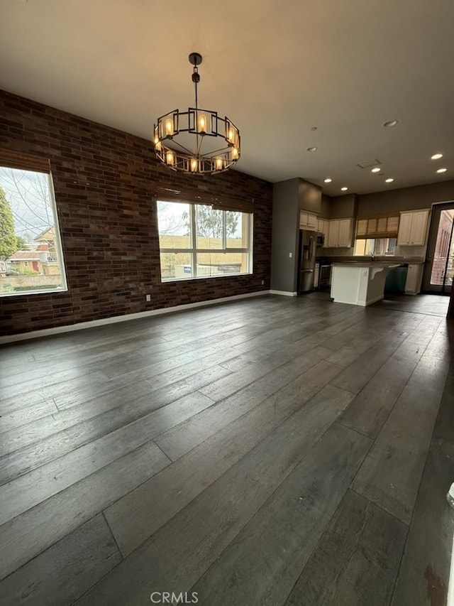 unfurnished living room with dark wood-type flooring, a chandelier, a healthy amount of sunlight, and brick wall