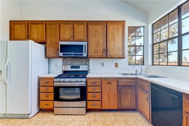 kitchen featuring tile countertops, lofted ceiling, appliances with stainless steel finishes, brown cabinets, and a sink