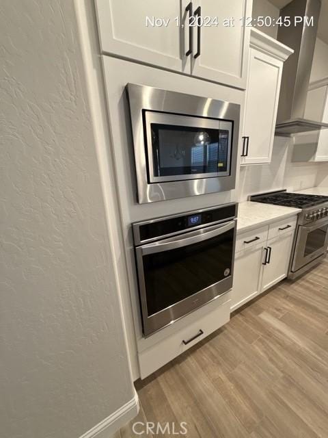 kitchen with a textured wall, appliances with stainless steel finishes, white cabinetry, wood finished floors, and wall chimney exhaust hood