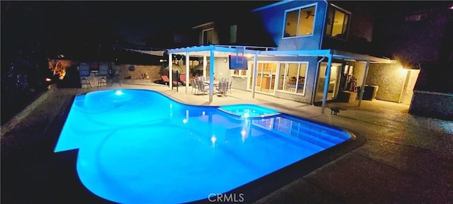 pool at night with a pool with connected hot tub, a patio area, and a pergola