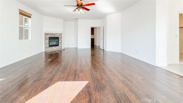 unfurnished living room with dark wood-style flooring, ceiling fan, a stone fireplace, and baseboards