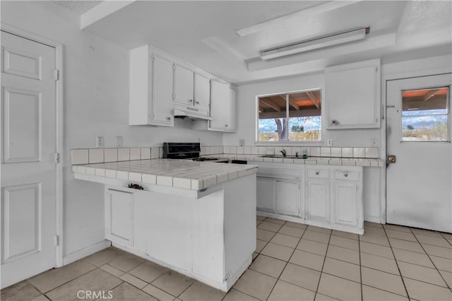 kitchen featuring tile countertops, under cabinet range hood, a peninsula, white cabinets, and range