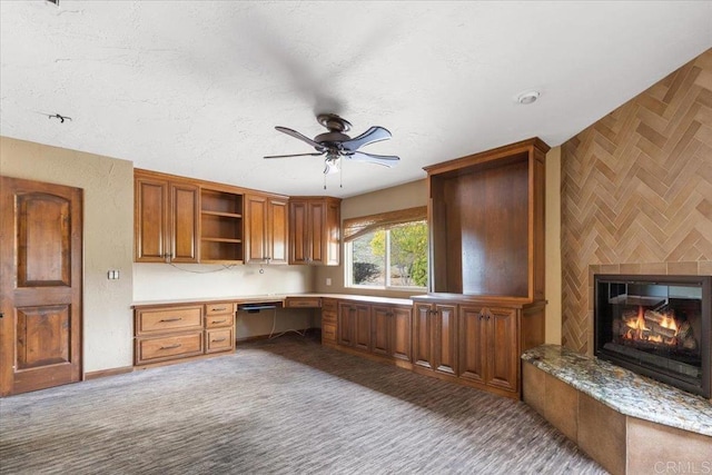 unfurnished office with a textured ceiling, carpet floors, a ceiling fan, built in desk, and a glass covered fireplace