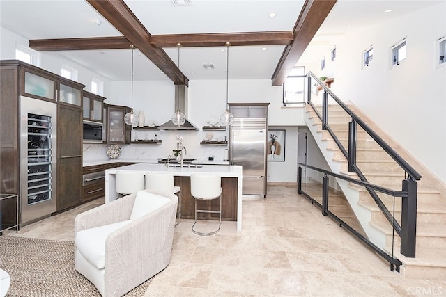 kitchen featuring stainless steel appliances, dark brown cabinetry, wall chimney range hood, and beamed ceiling