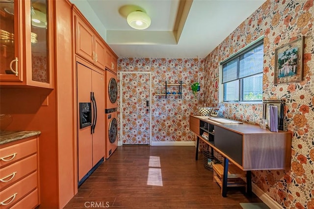 kitchen featuring dark wood-type flooring, stacked washer / drying machine, wallpapered walls, glass insert cabinets, and baseboards