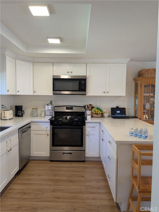 kitchen with a raised ceiling, white cabinets, appliances with stainless steel finishes, a peninsula, and light wood-type flooring