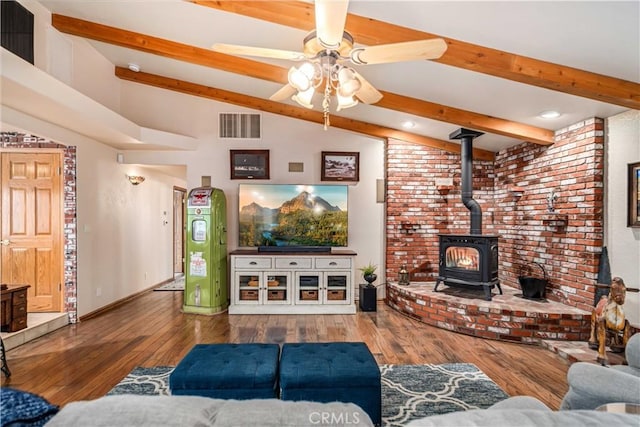 living area with visible vents, a ceiling fan, wood-type flooring, a wood stove, and vaulted ceiling with beams